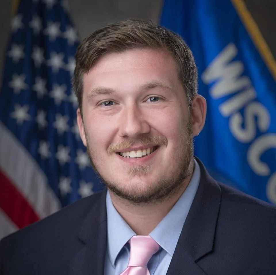 Portrait of a young man in a suit smiling, standing in front of American flags.