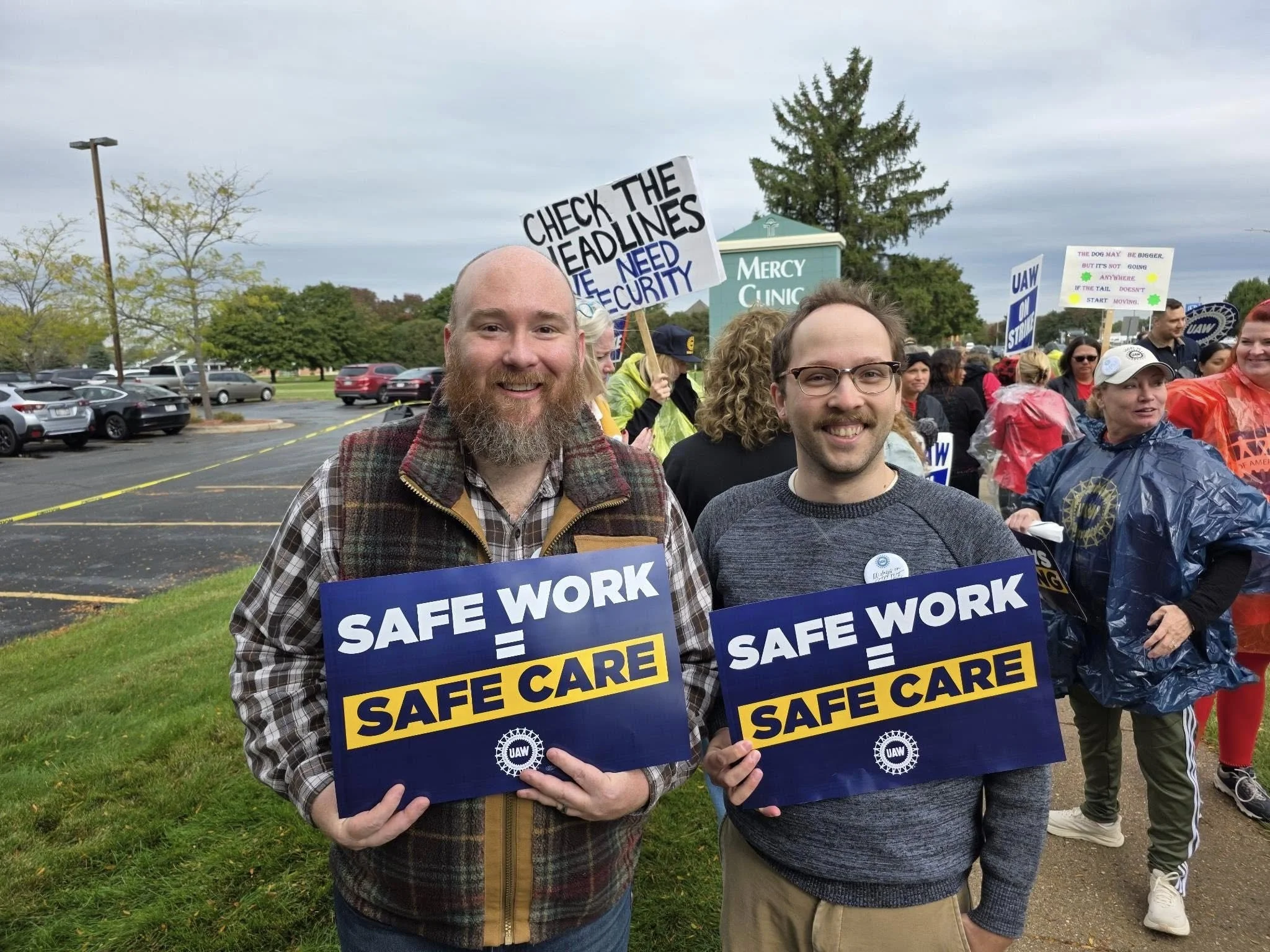 Two men smiling and holding signs that read 'SAFE WORK = SAFE CARE' at a protest or rally with other people holding signs in the background.