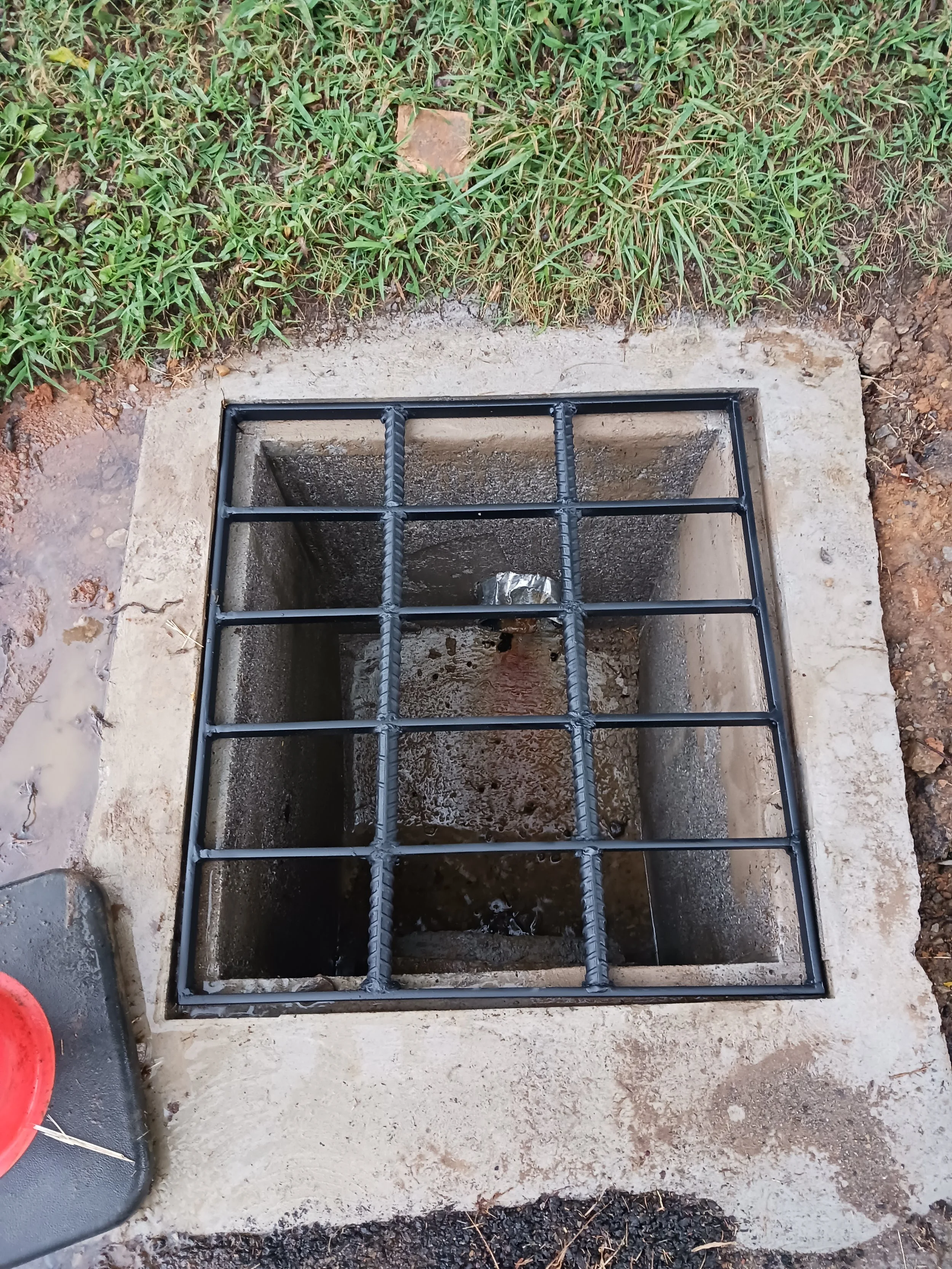 A square manhole cover with a grid of black metal bars on top, surrounding a concrete pit with water at the bottom, positioned in a grassy outdoor area.