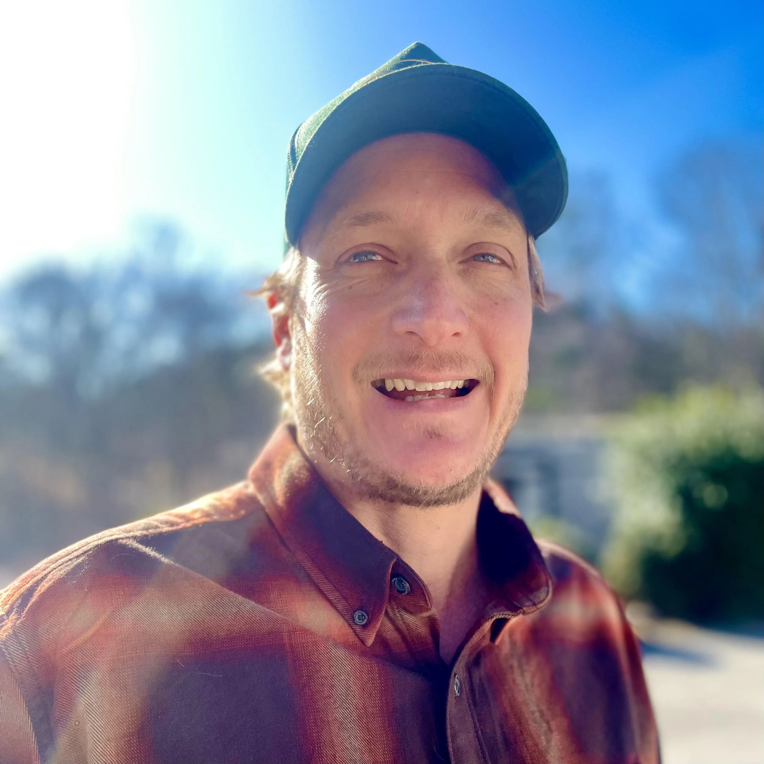 Portrait of a smiling man wearing a baseball cap and a plaid shirt outdoors on a sunny day.