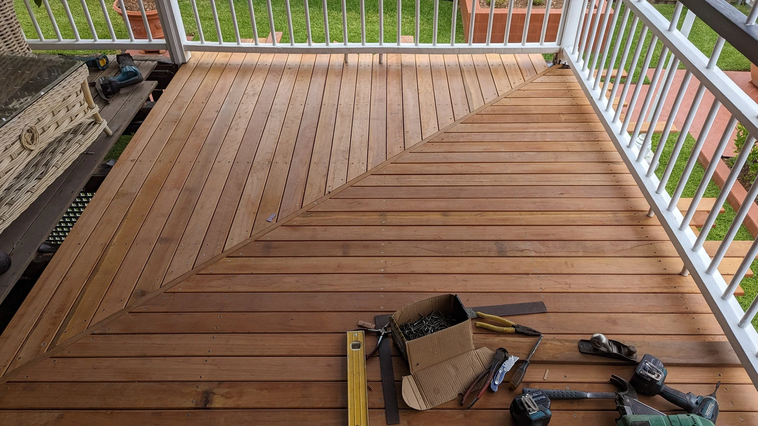 Freshly installed wooden porch flooring with tools and hardware scattered around, white railing, a wicker chair, and a view of a grassy yard.