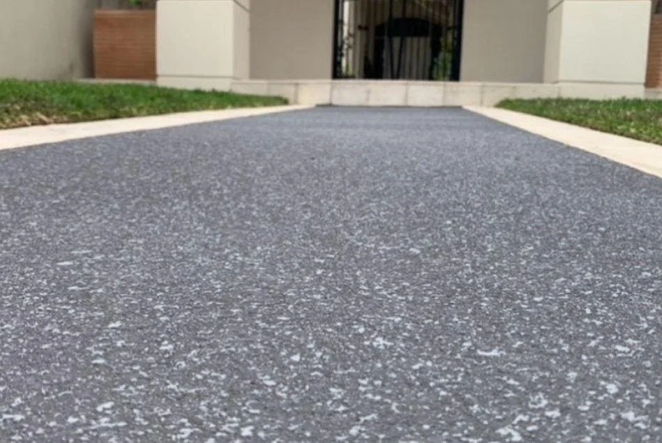 Close-up of a speckled gray asphalt driveway leading to the entrance of a modern house with a black gate and concrete steps.