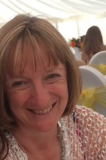 A smiling woman with shoulder-length brown hair in a lacy white top, in an indoor event setting with decorated tables and other guests.