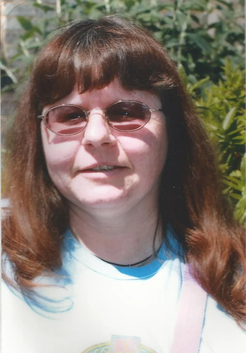 A woman with red hair wearing sunglasses and a white shirt outdoors with green foliage in the background.