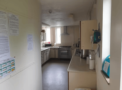 Empty kitchen with white cabinets, stove, and countertop, with a small window and some notices posted on the wall.