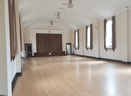 Empty room with wooden floor, white walls, large windows with dark curtains, and a small stage at the front.