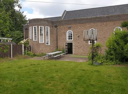 A brick building with large bay windows, a basketball hoop, and a picnic table in a grassy yard.