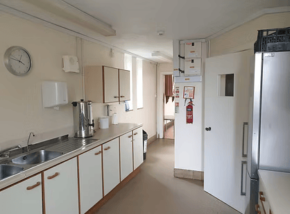 Kitchen with white cabinets, stainless steel sink, wall clock, and refrigerator, with a fire extinguisher and boxes on the wall near a door.