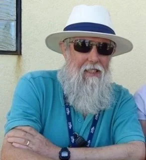 Older man with a white beard wearing sunglasses, a white hat with a blue band, and a blue shirt, standing outdoors.