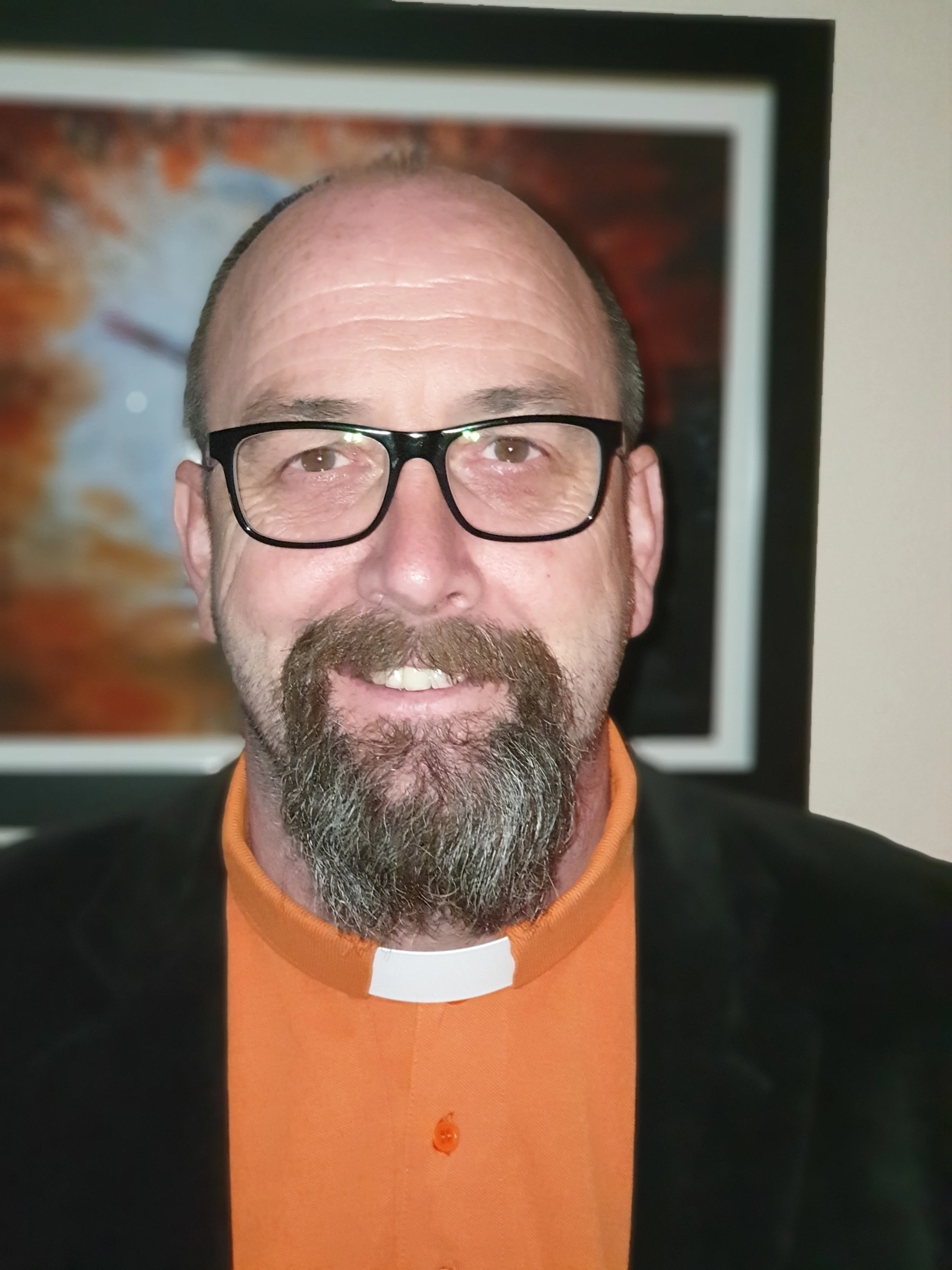 A middle-aged man with glasses, a beard and mustache, wearing an orange clerical shirt with a white collar and a black jacket, smiling in front of a framed colorful abstract artwork.