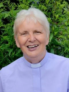 An elderly woman with short white hair, smiling, wearing a light purple clergy shirt, standing outdoors in front of green foliage.