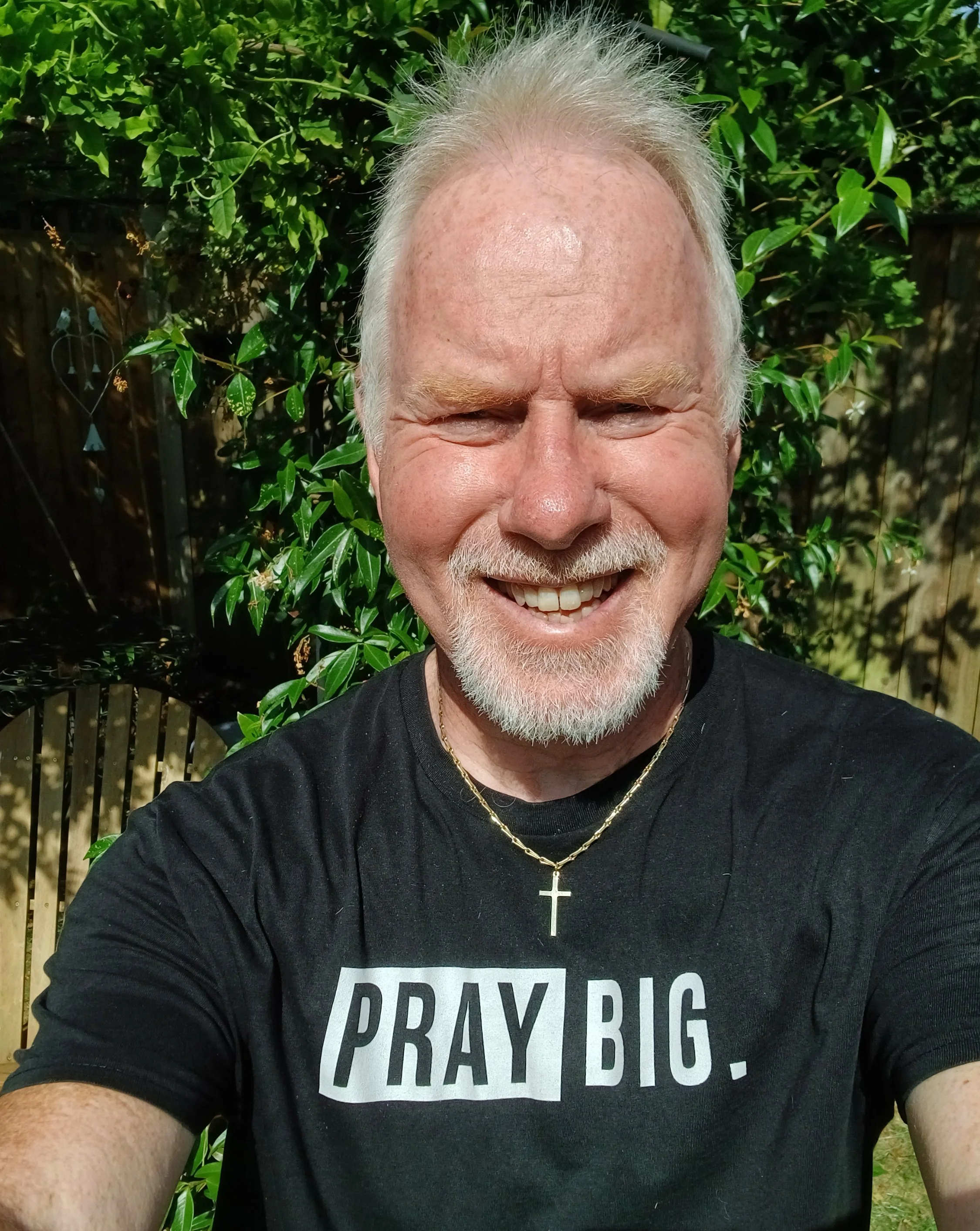 A smiling man with white hair and a beard taking a selfie outdoors, wearing a black T-shirt with the words 'PRAY BIG.' and a gold cross necklace, with greenery and a wooden fence in the background.