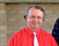 A man with gray hair smiling, wearing a red jacket and a white shirt with a red striped tie, standing outdoors in front of a beige brick wall.