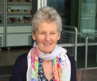 An elderly woman with short gray hair smiling, wearing a colorful scarf and a dark top, standing outside a store.