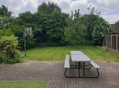 A backyard with a brick patio, a picnic table, a grassy area, a basketball hoop, and green trees and bushes.
