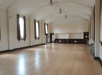 Empty large room with wooden floors, high ceiling, ceiling fans, four tall windows with curtains, and built-in cabinets or counters at the front.