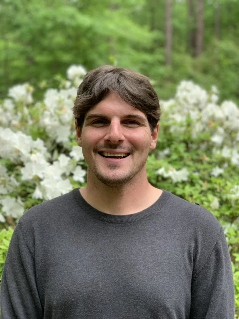A smiling man with dark brown hair and a stubble, wearing a dark gray sweater, standing outdoors in front of a lush green background with white flowers.