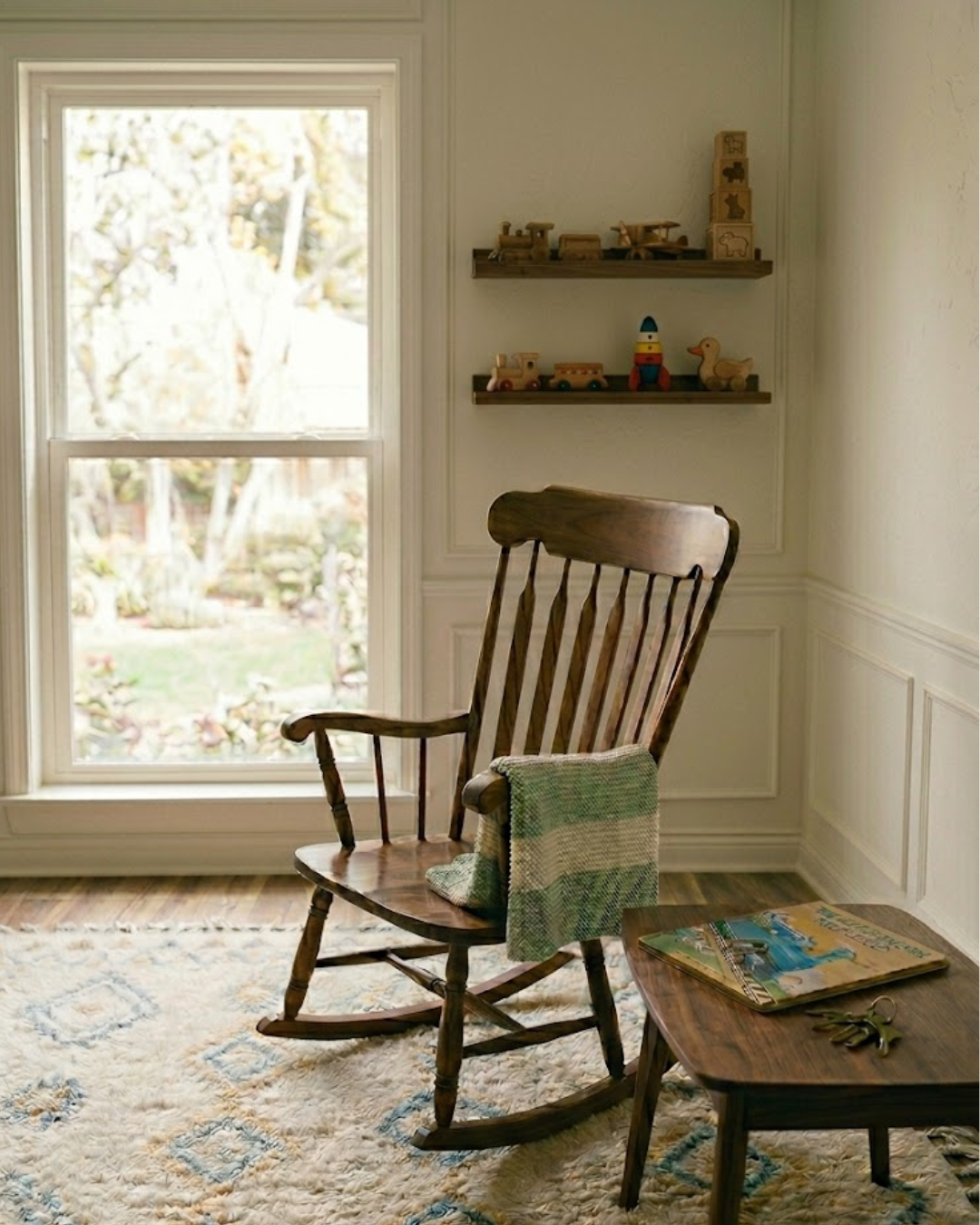 A cozy room with a wooden rocking chair placed next to a window, a small wooden table with a magazine and a set of keys, and two shelves on the wall with wooden toys and puzzles. Outside, trees and a garden can be seen.