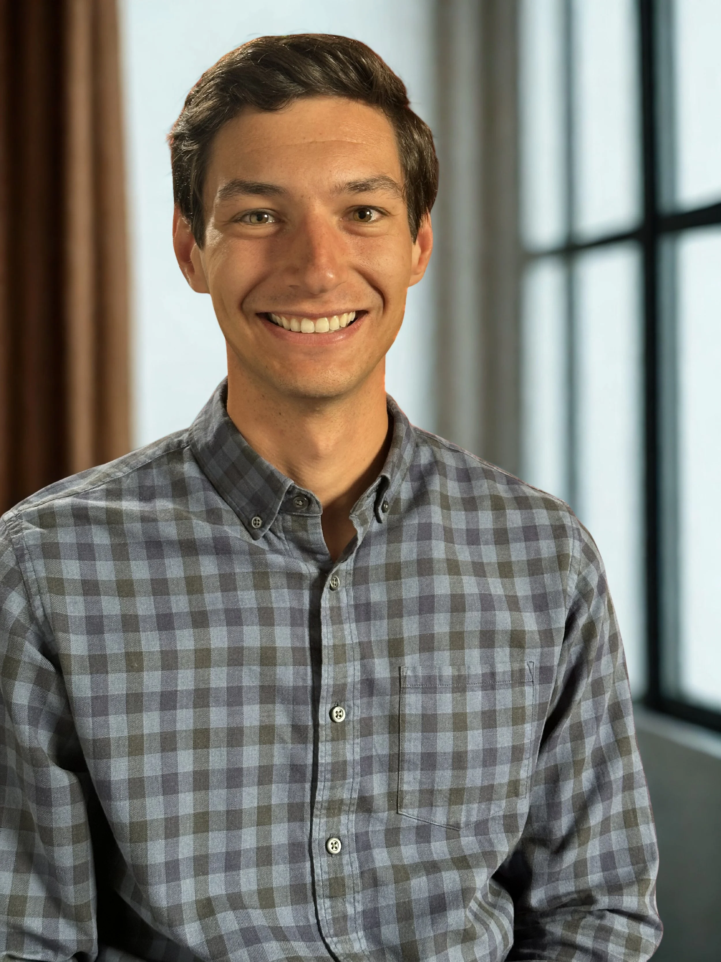 A young man with dark brown hair, wearing a checkered button-up shirt, smiling in front of a window with curtains and natural light.