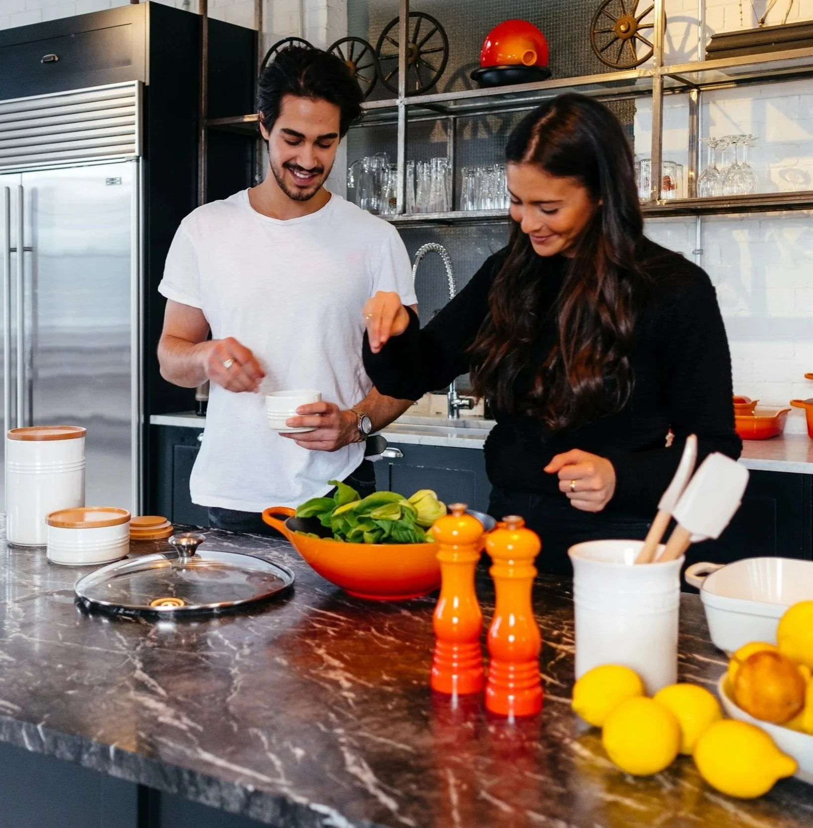 Two people stand in a modern kitchen, smiling and possibly preparing a meal together. The man holds a bowl, and the woman is reaching into it. There are various kitchen items on a dark marble countertop, including a large orange bowl filled with leafy greens, lemons, orange salt and pepper shakers, and several white and orange containers. Glassware and shelves are visible in the background.