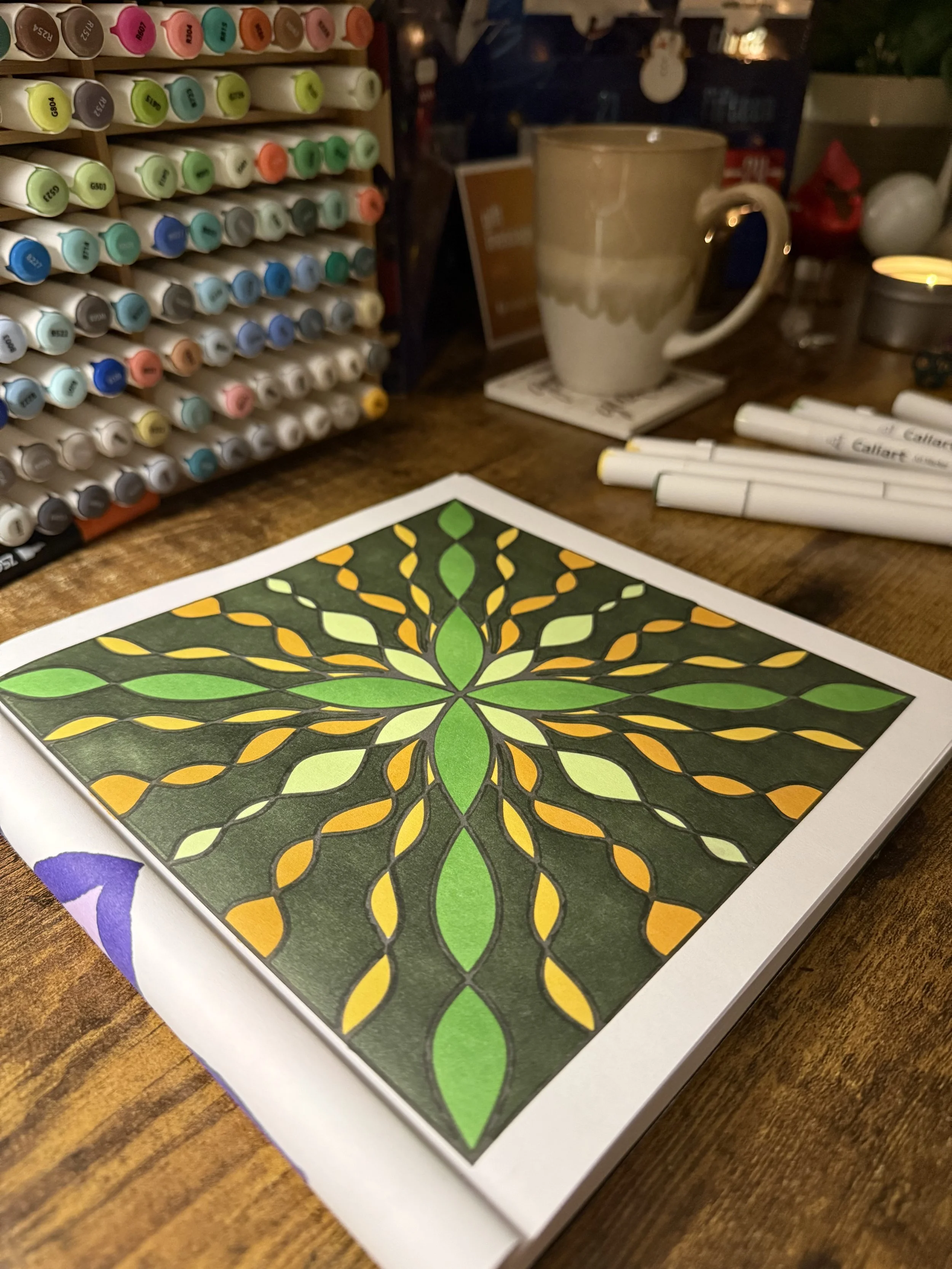 Hand-colored symmetrical design from Mindful Trine coloring book with markers, candlelight, and tea in a warm creative space