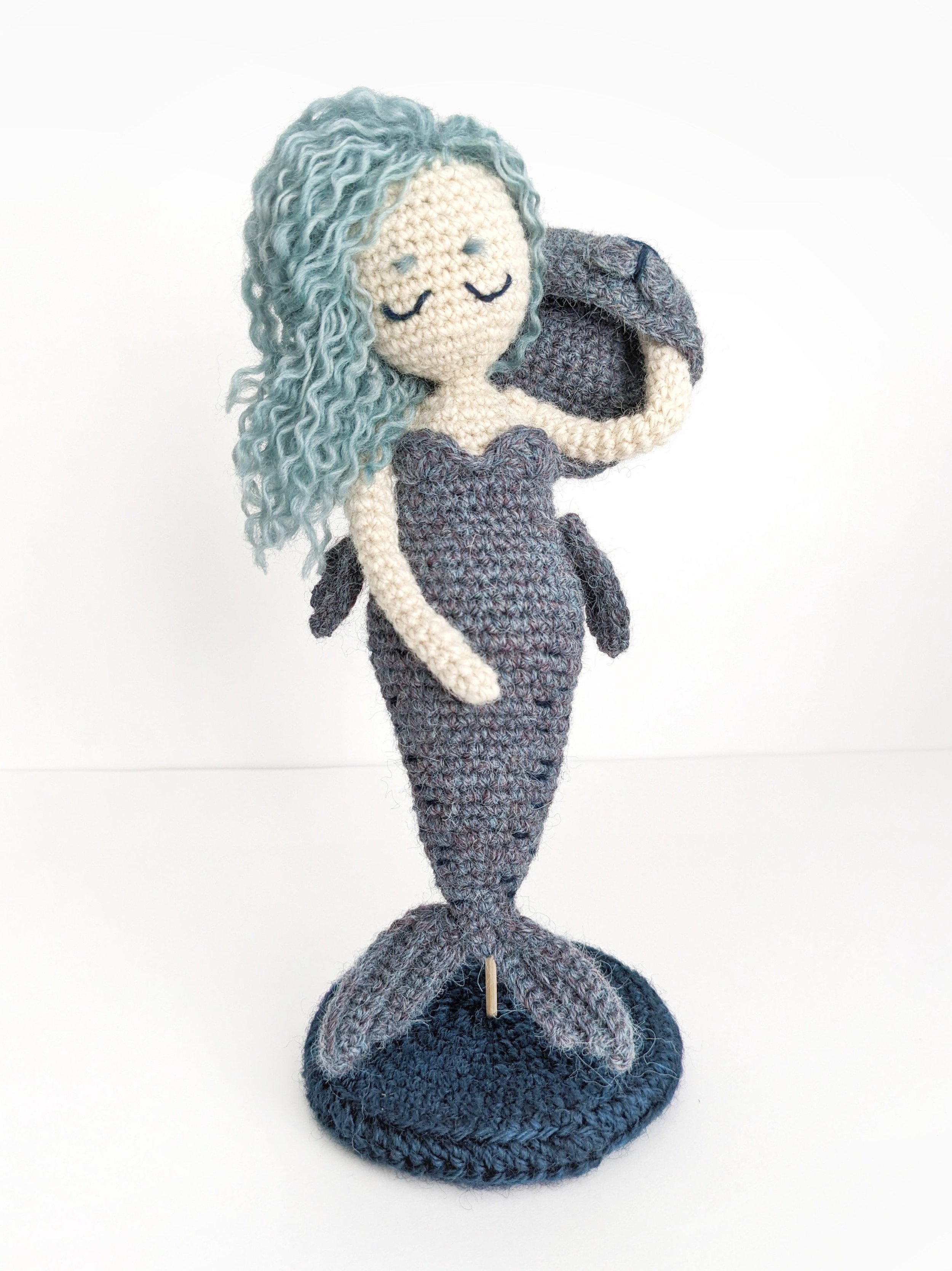 Crocheted mermaid doll with long, wavy blue hair, wearing a dark purple top and sitting on a round, dark blue crocheted base.
