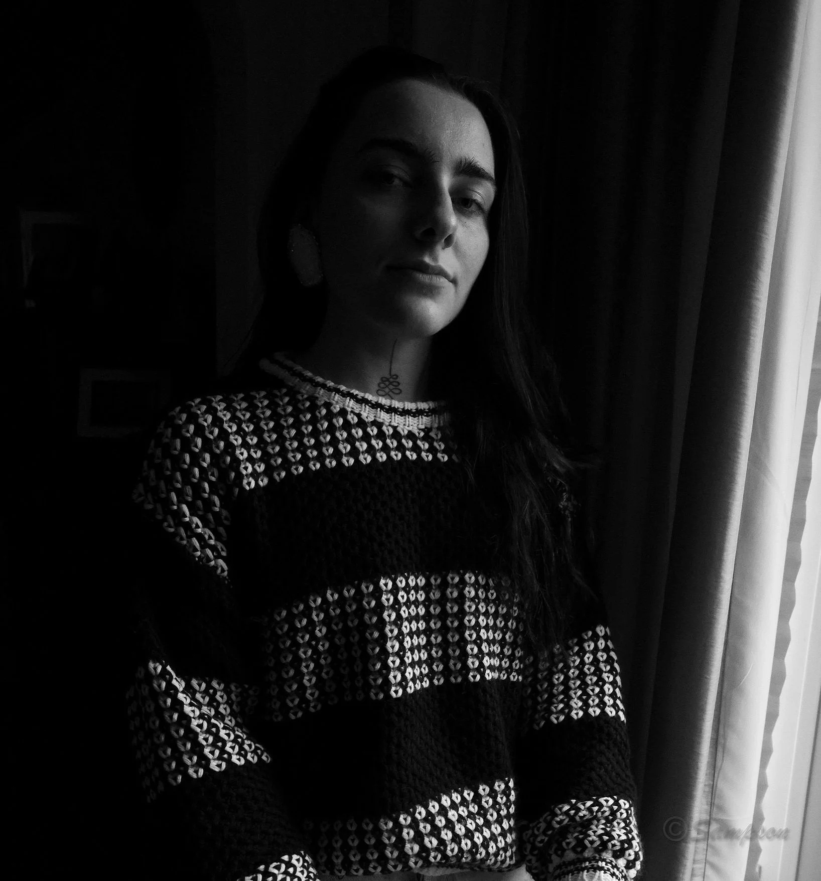Black and white photo of a person wearing a patterned sweater, standing near a window with curtains.