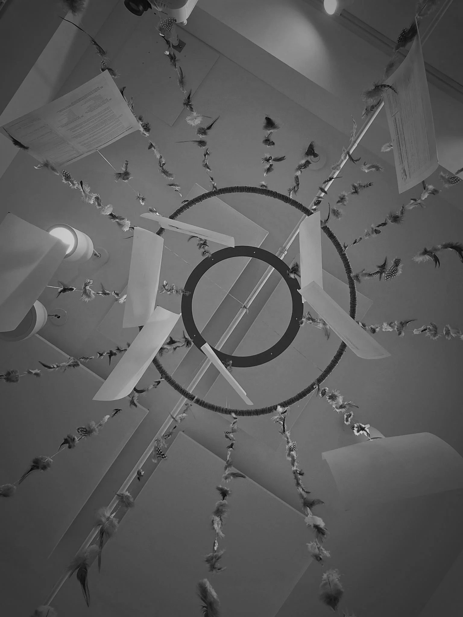 Black and white installation art viewed from below, featuring a circular frame with dangling feathers and papers suspended from the ceiling.