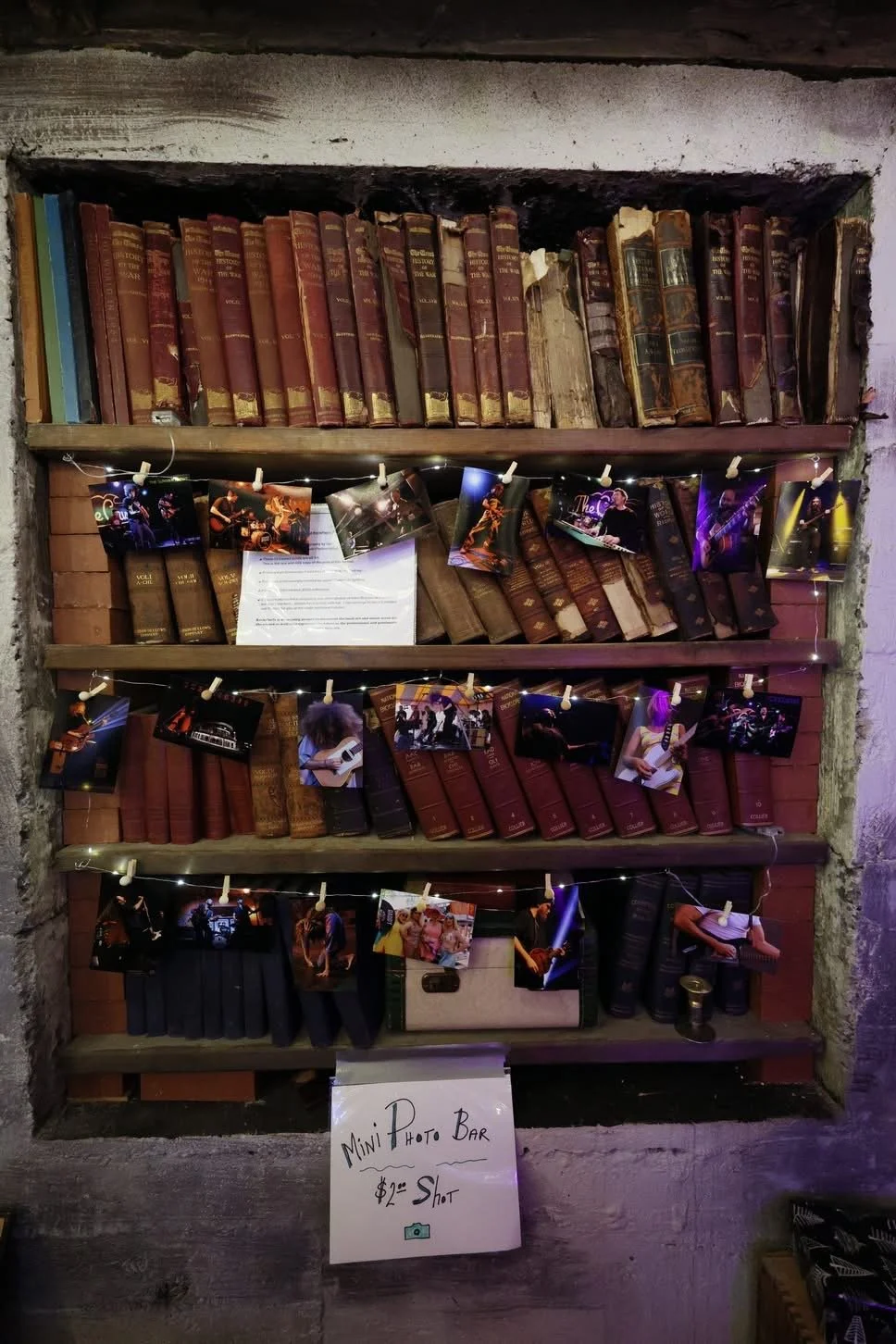 Bookshelf decorated with photos and fairy lights, with a sign reading 'Mini Photo Bar $2 a Shot' in front.