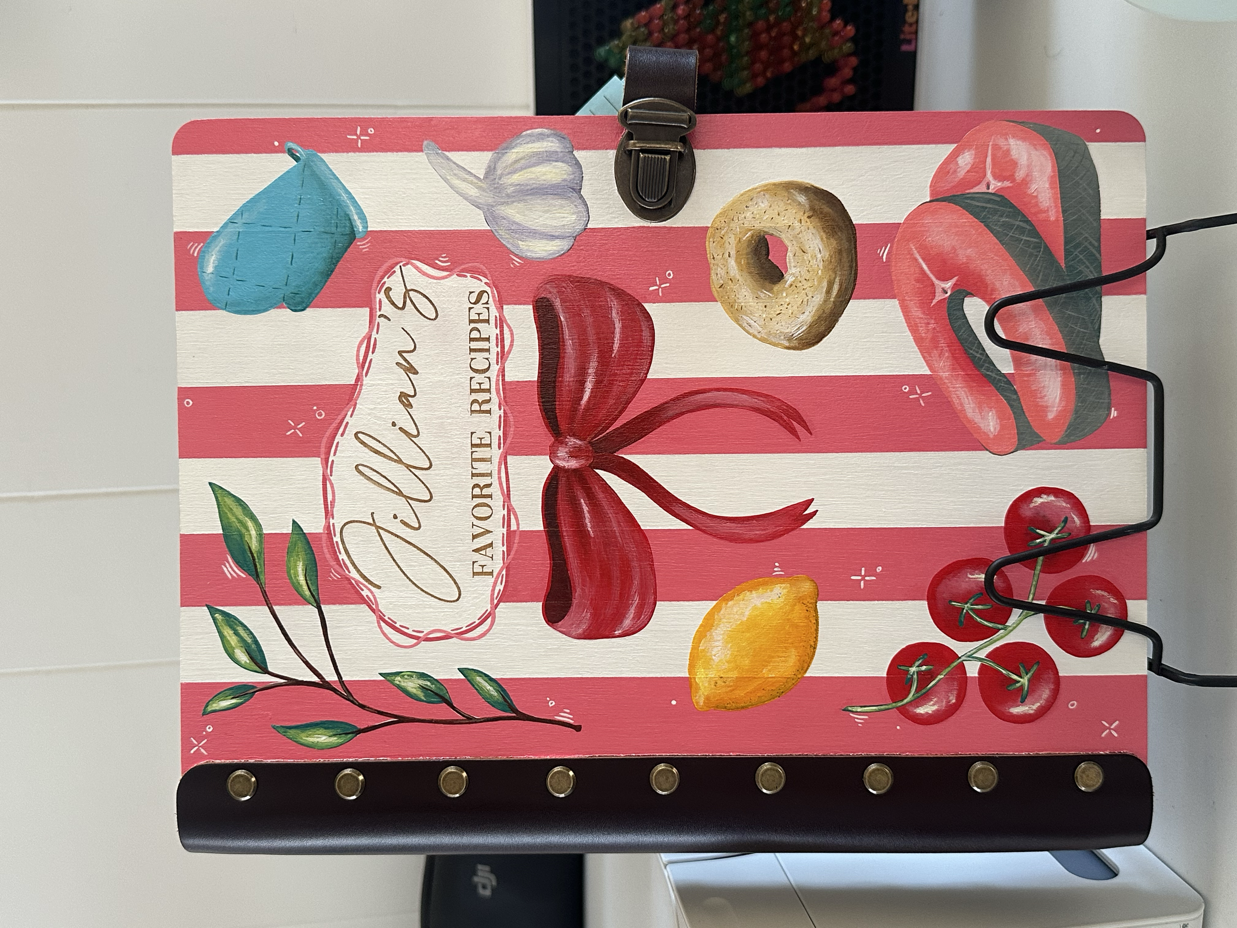 Decorative cookbook with red and white stripes, featuring illustrations of a blue oven mitt, garlic, a donut, tomatoes on the vine, a lemon, and a red bow, with a phrase "Jillian's Favourite Recipes".