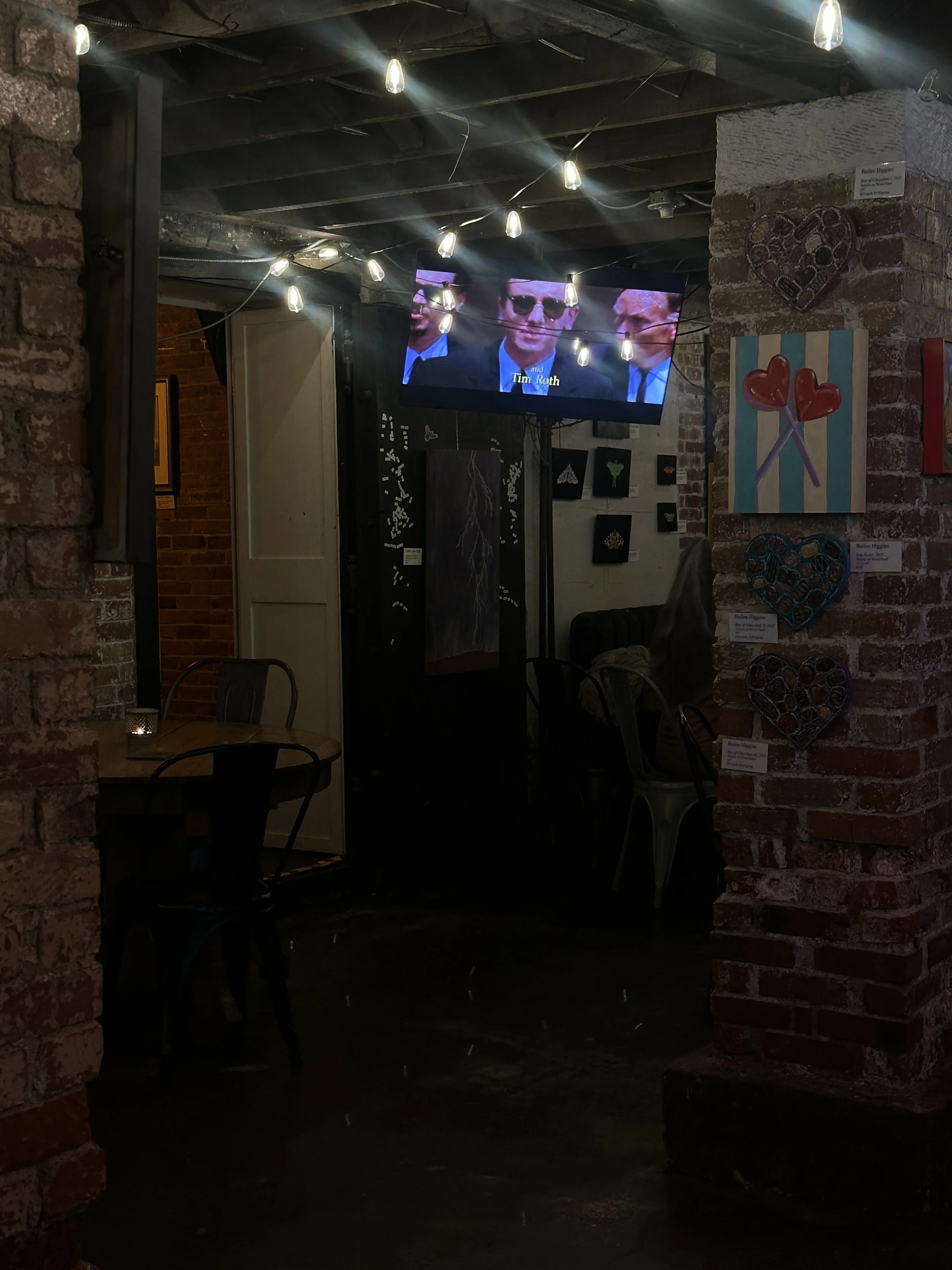 A dimly-lit bar, exposed brick walls and string lights hanging from the ceiling. There is a television mounted on the wall displaying a scene with men in suits and sunglasses. On the right wall, there are colorful heart-shaped and lollipop artwork.