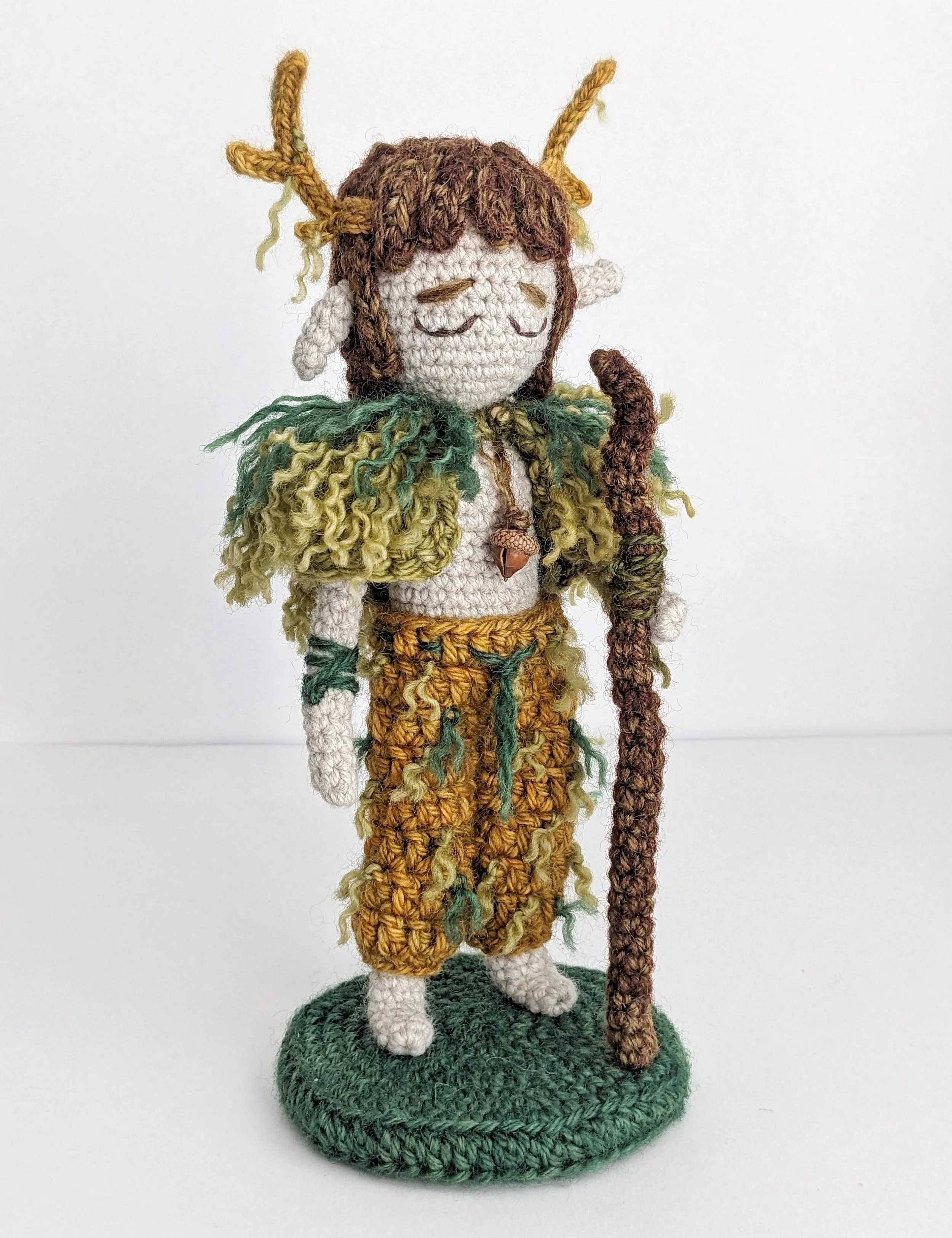 Crocheted doll resembling a mythological forest creature with antlers, closed eyes, and a beard, standing on a green base.