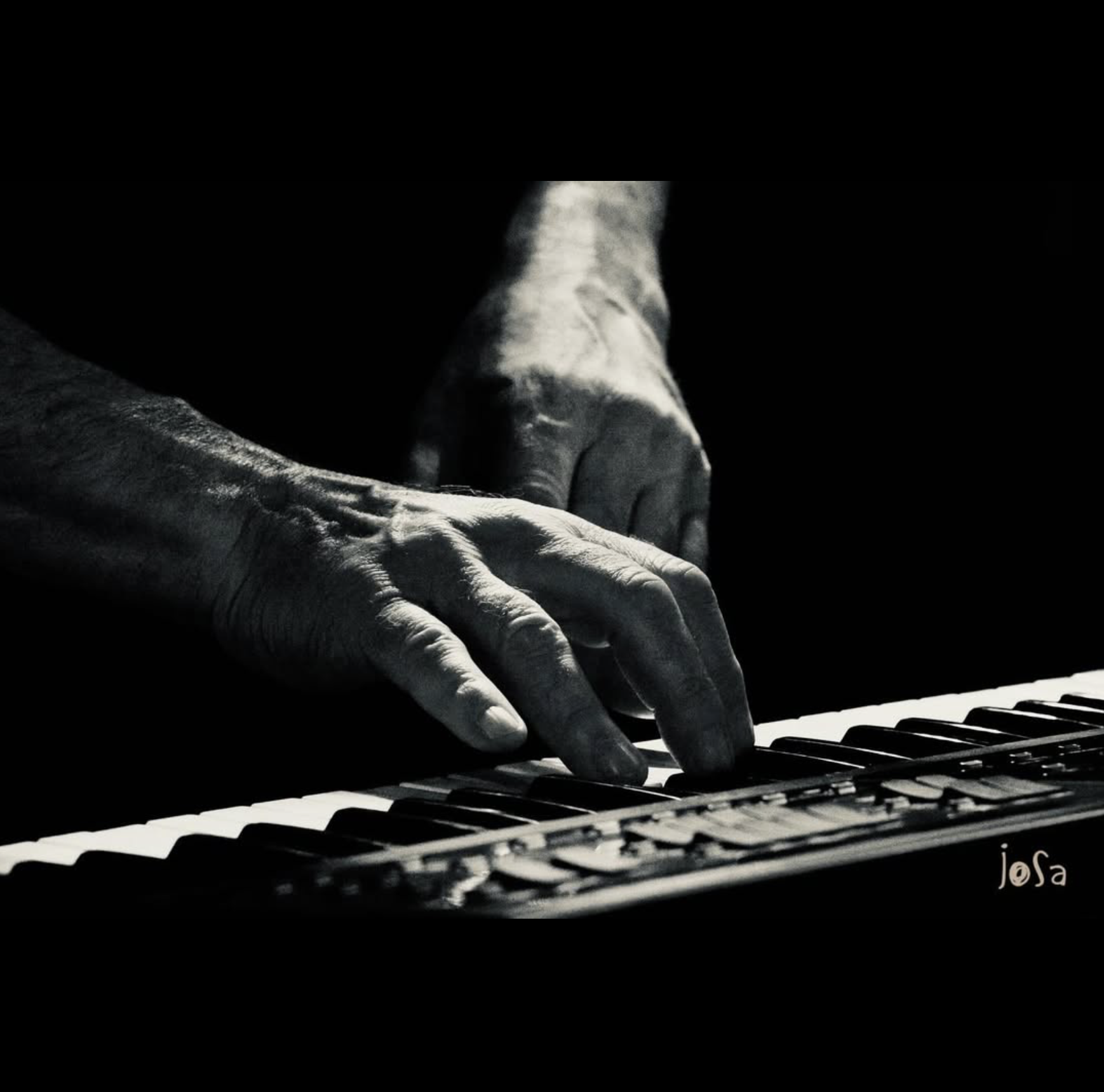Black and white photograph of hands playing a piano, with the focus on the fingers pressing keys, signed by Josa in the bottom right corner.