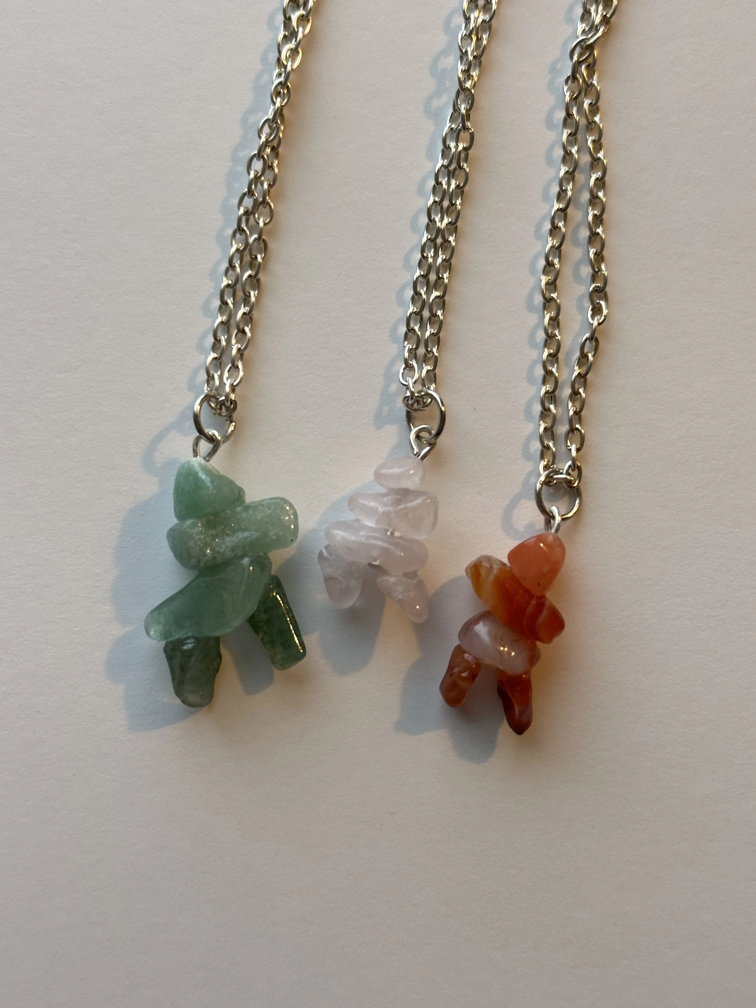 Three Inukshuk necklaces each with a different colored stone pendant hanging on a silver chain, positioned on a white background.