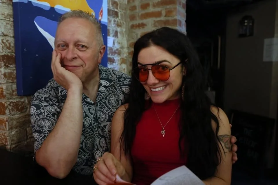 Two people smiling and sitting close together in a cozy, brick-walled café or restaurant. The older man has short blonde hair and is wearing a patterned shirt. The young woman has long black hair, orange tinted sunglasses, and is wearing a red sleeveless top with jewelry.