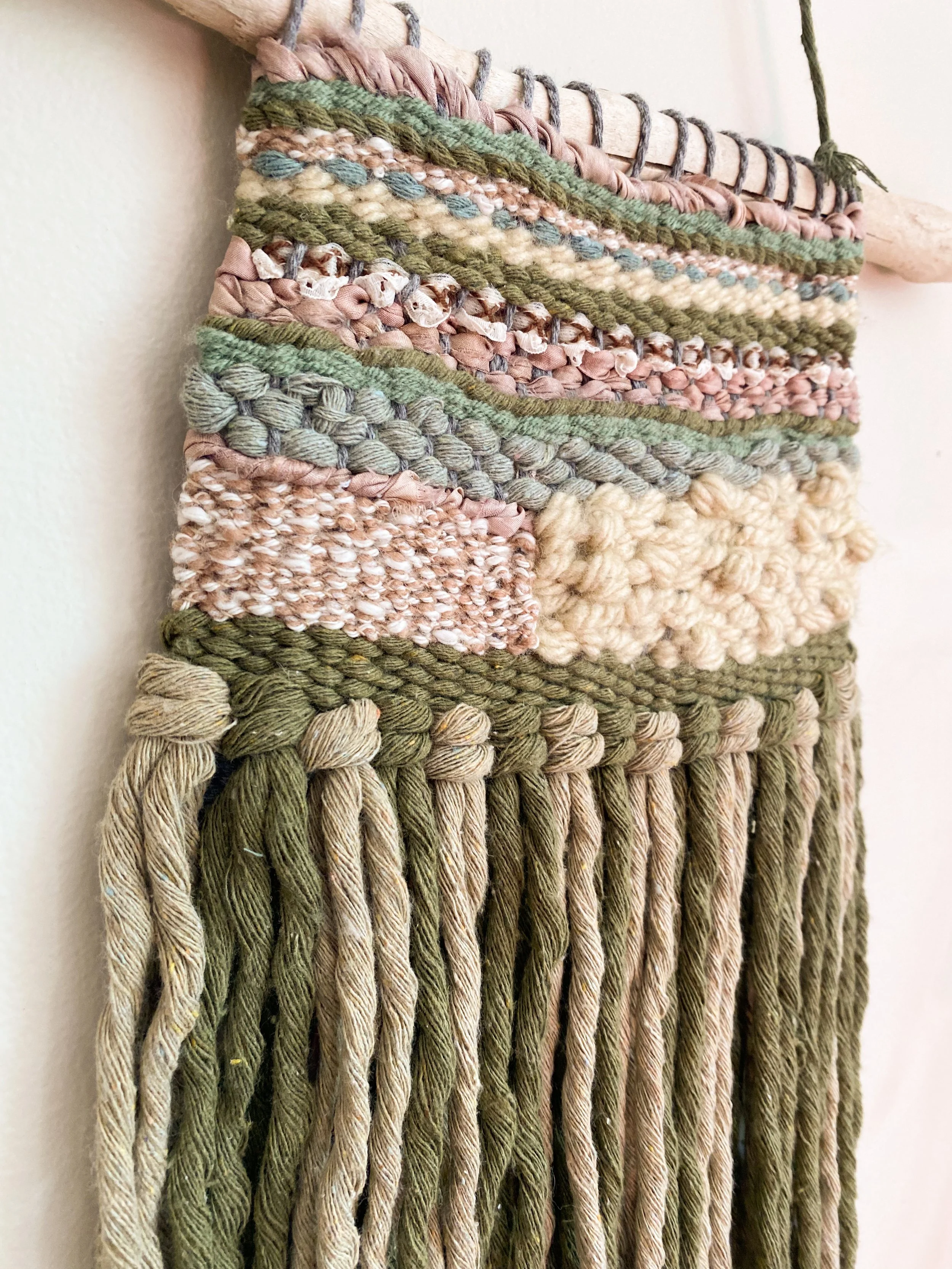 Close-up of a woven wall hanging made from various textured yarns in earth tones, including greens, beiges, and pinks, with fringe at the bottom.