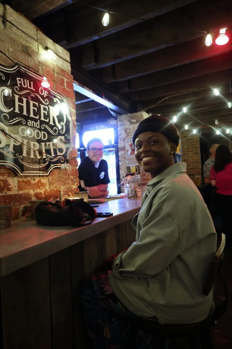 A woman sitting at a bar with a smile, wearing a beige jacket and a black beanie. There is a man behind the bar smiling, and the bar has a sign that reads 'Full of Cheer and Good Spirits.'