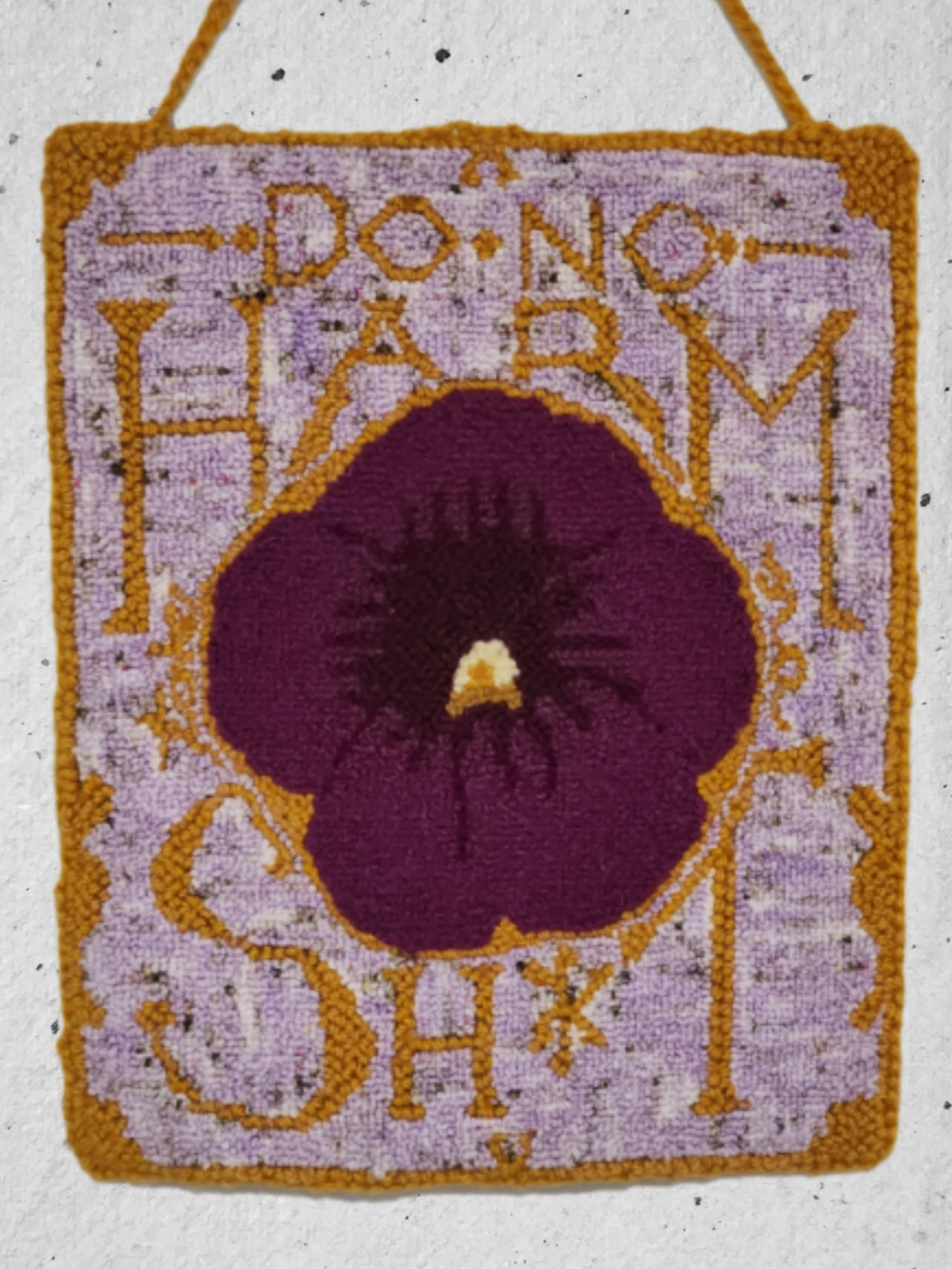 A small decorative rug with a purple background, a central design of a steeple-like shape in yellow, and surrounded by yellow patterns; the rug has a yellow border and a loop at the top for hanging.
