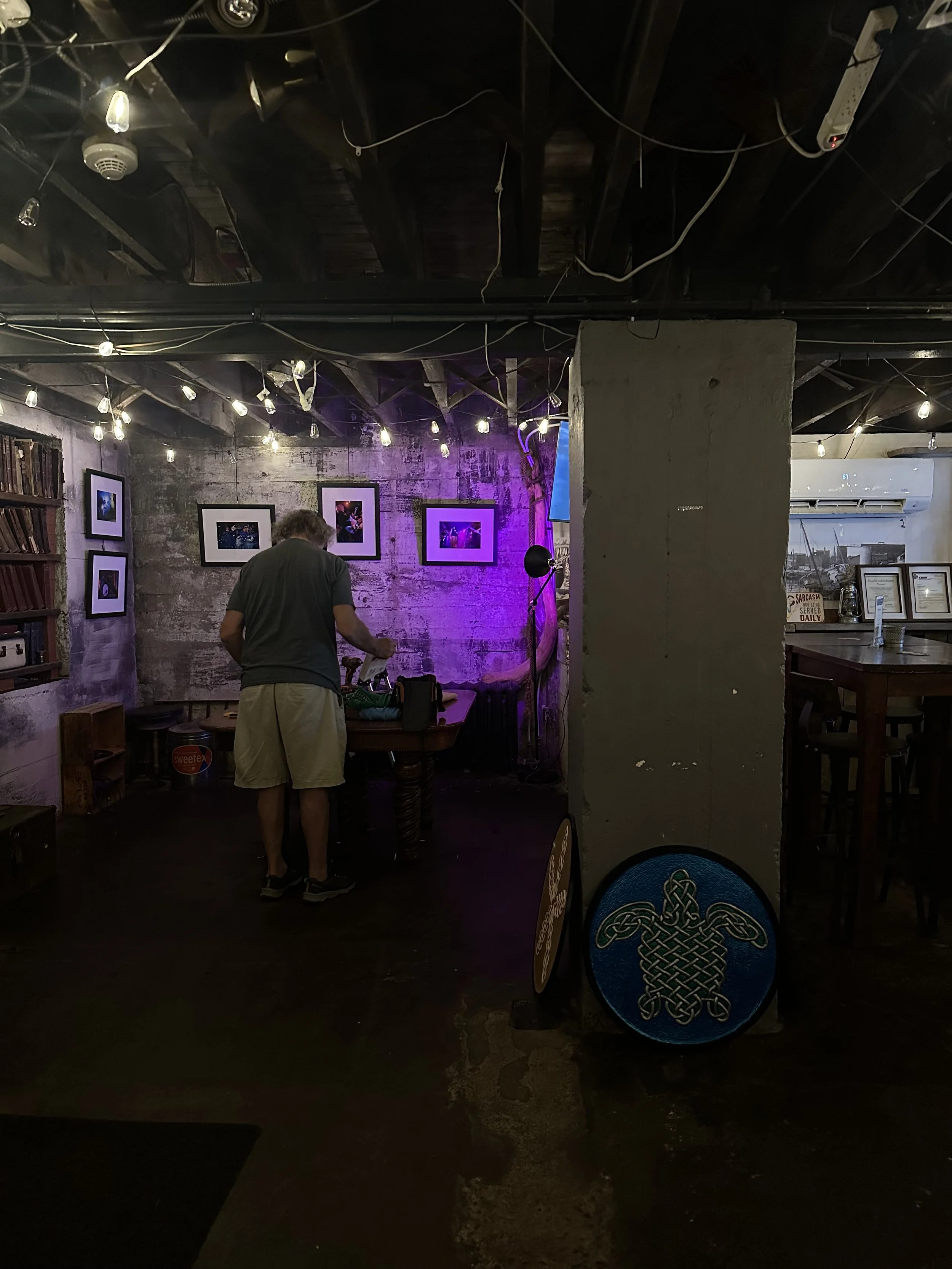 A person standing near a table in a dimly lit room with string lights and framed pictures on the wall, featuring a concrete pillar with a decorative round sign.