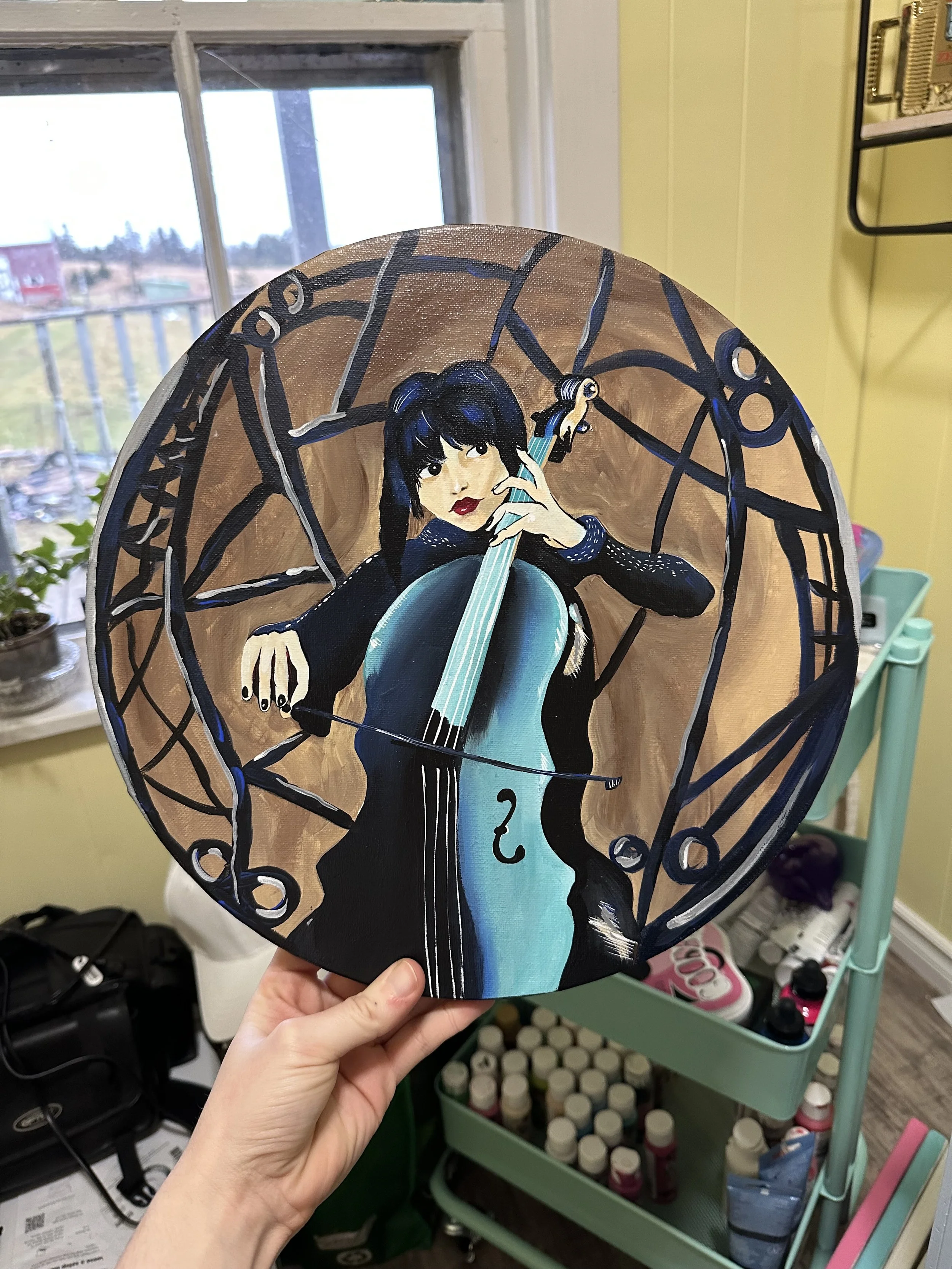A hand holding a circular painted artwork of wednesday addams, with black hair playing a cello, set against an abstract background with a spherical design.