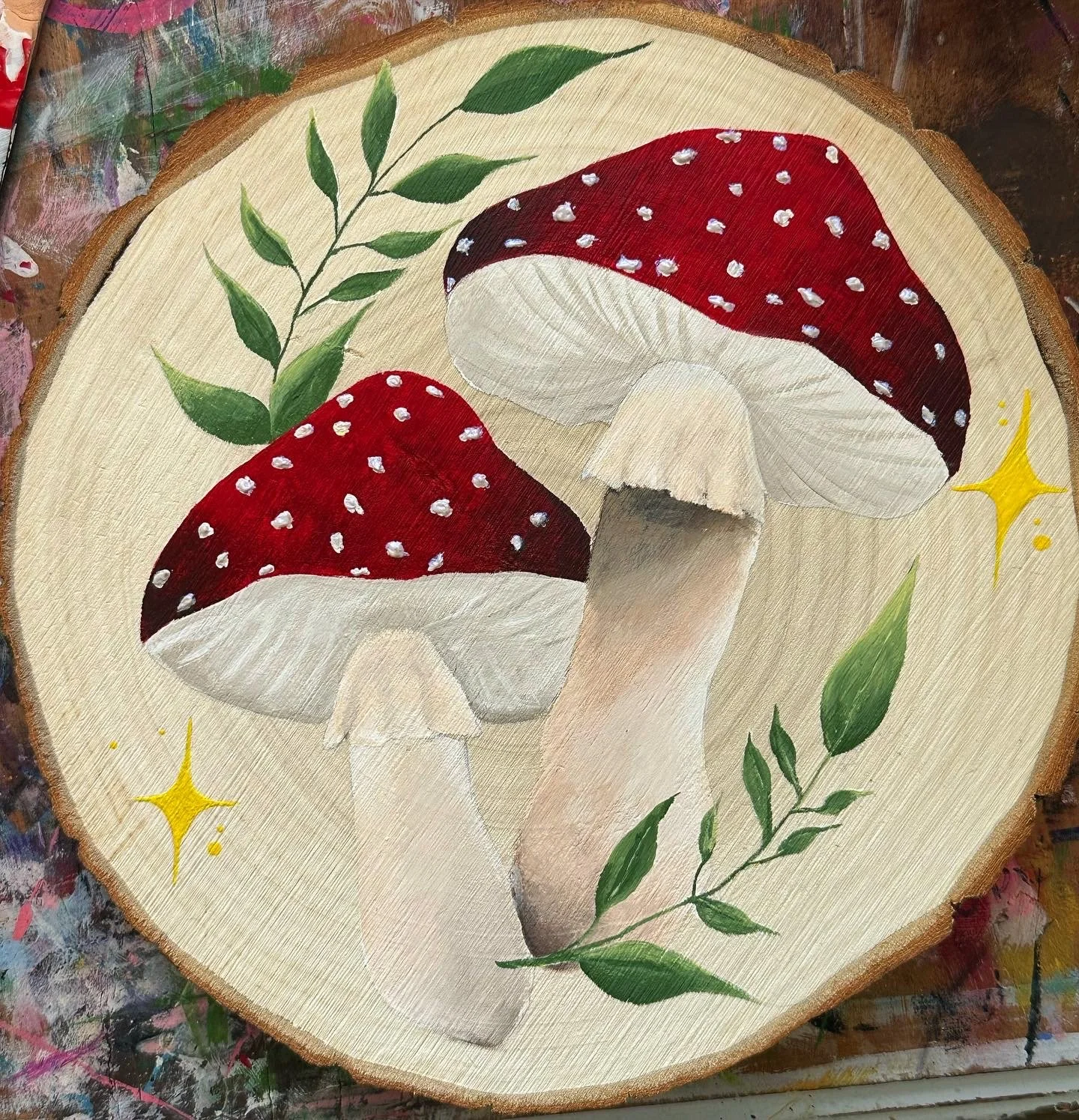 Painting of two red and white mushrooms surrounded by green leaves and yellow star-like accents on a circular wooden surface.