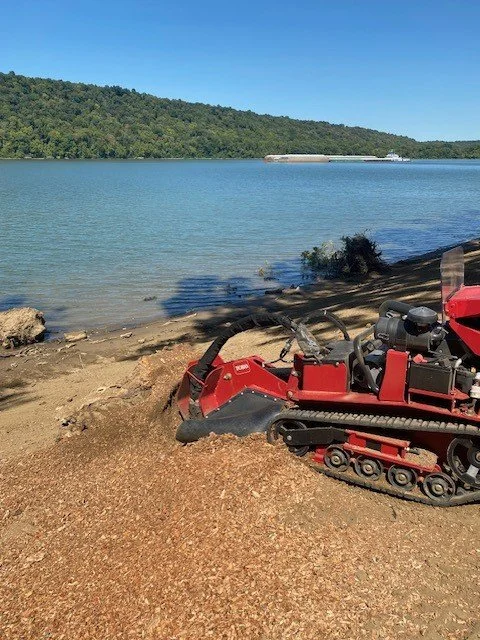 A red snowcat tracked vehicle parked on a dirt shore next to a body of water with green hills across the water and a clear blue sky.