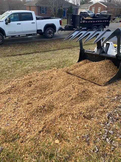 A construction site with a large pile of dirt and a backhoe excavator, with a white pickup truck and a black trailer in the background.