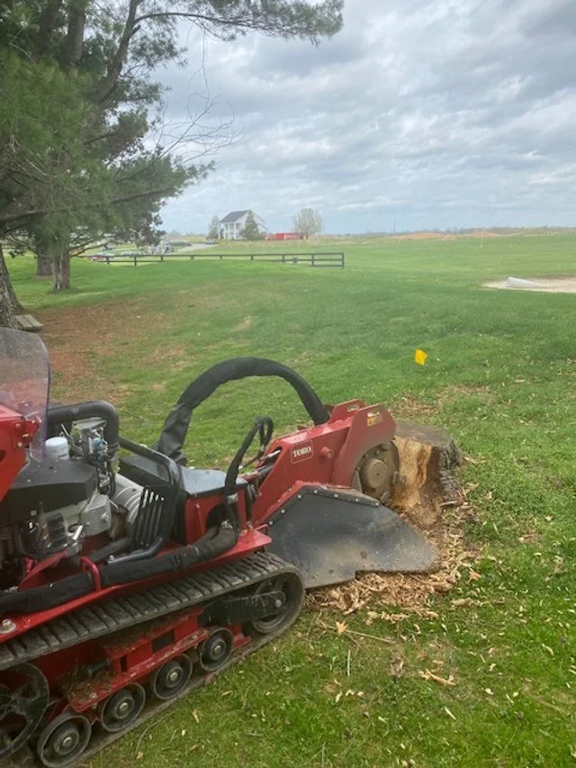 A tracked wood cutting machine with a large spinning saw blade on a grassy field, with a tree and a house in the background.