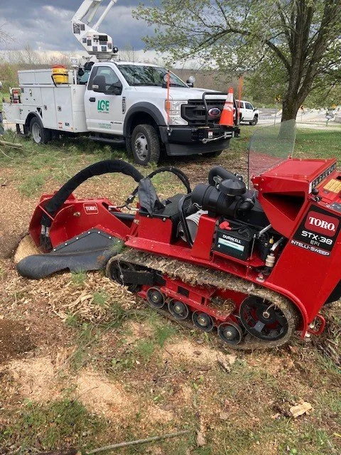 A red Toro ground grinder with tracks is parked on a patch of grass and dirt, with a white utility truck equipped with a bucket lift and traffic cones in the background.