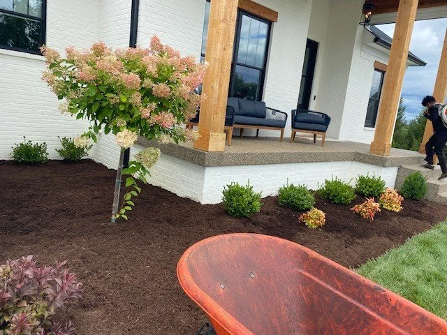 Front porch of a house with wooden support beams, black outdoor furniture, and a landscaped garden with flowering bushes and mulch.