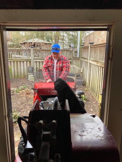 Person in a red plaid shirt and blue cap is operating a red lawnmower outside in a backyard, viewed from inside a house through a window with an iron railing partially blocking the view.