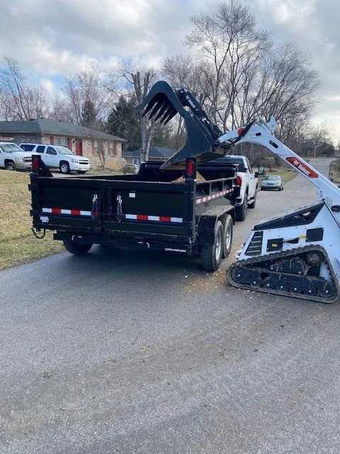 A black flatbed truck with a white mini excavator attached to its bed, parked on a residential street with houses and cars in the background.