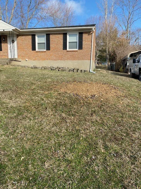 Front view of a brick house with two windows, black shutters, and a small front porch, with a lawn and a few leafless trees in the background, and a white vehicle parked on the driveway to the right.