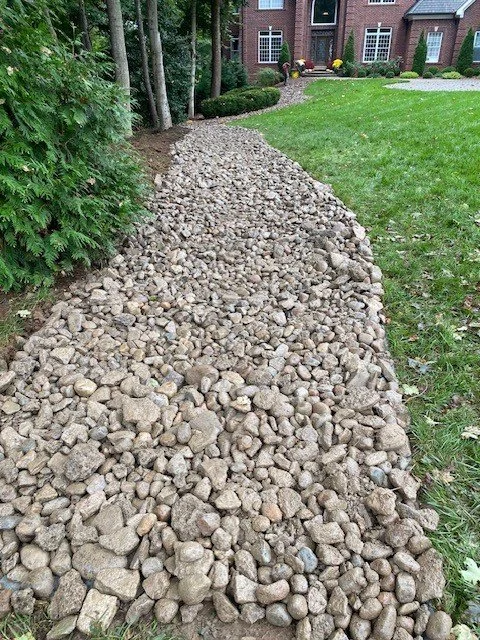 Rock pathway alongside a grassy yard with shrubs and trees in front of brick houses.